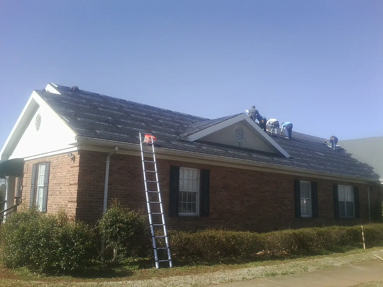 Crew tearing shingles off a brick ranch roof with a ladder set — Metal Roof Repair work by our crew in Mill Spring